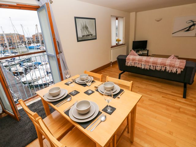 A dining table set for four next to an open balcony door overlooking boats and a living room with a sofa and a television at 15 Sovereign House in Milford Haven