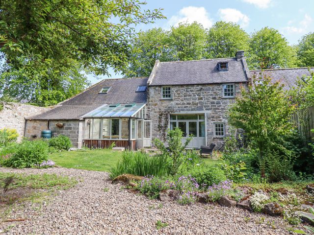A stone house with a glass conservatory and garden with plants and gravel path at The Old Coach House in Eglingham near Alnwick