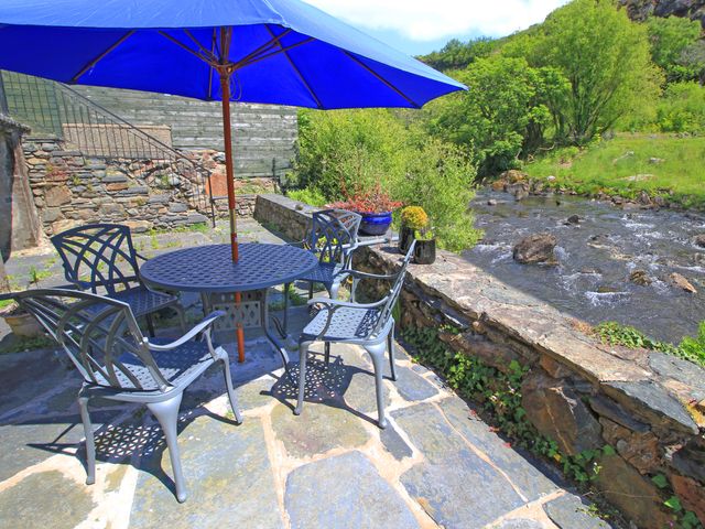 An outdoor patio with metal chairs and a table under a blue umbrella next to a river at Riverbank Cottage in Beddgelert