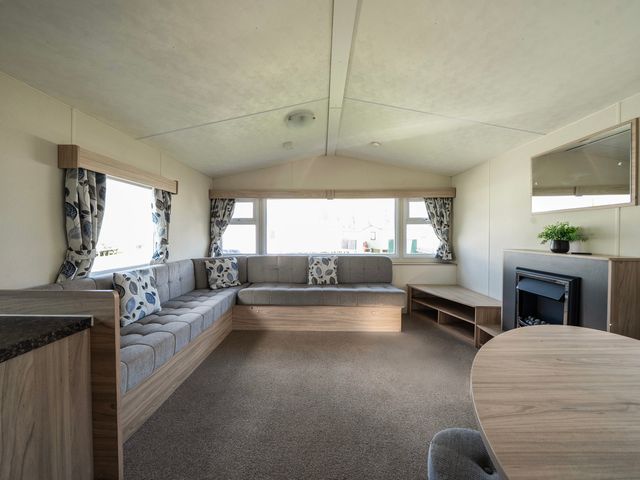 A living room with an L-shaped sofa cushions a round table a fireplace and windows with curtains at Ripples & Reeds - Billing Aquadrome in Billing Aquadrome Holiday Park