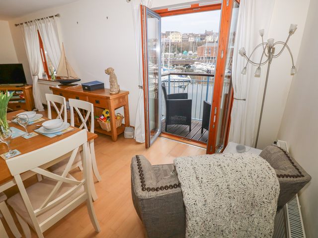 A dining area with set table and chairs near a sitting area with an armchair a side table and a floor lamp next to open doors leading to a balcony with two chairs overlooking a marina at 14 Neptune House in Milford Haven