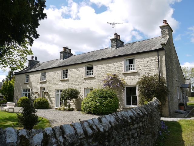 A stone cottage with multiple chimneys surrounded by bushes and a stone fence at 1 The Green in Silverdale