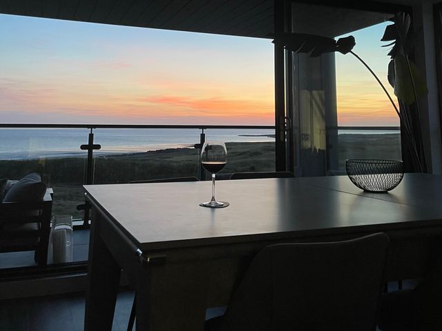 A dining table with a glass of wine and a decorative bowl near a window overlooking the sea at sunset at The Links 40 An apartment with spectacular sea views Porthcawl