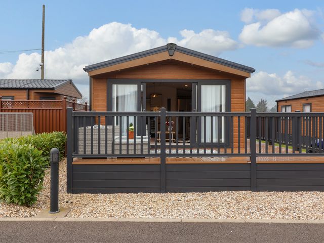 A wooden lodge with glass doors and a black railing on a deck at Lodge 36 - Kingfisher Lodge in Wickhamford near Evesham