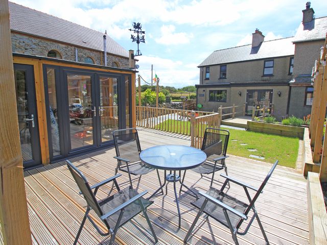 An outdoor wooden deck with a round glass table and four black chairs near a lawn and houses at Shooting Star Cottage in Pentrefelin near Criccieth