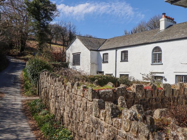 A white cottage with multiple windows behind a stone wall next to a narrow road at 2 New Mill Cottages in North Bovey