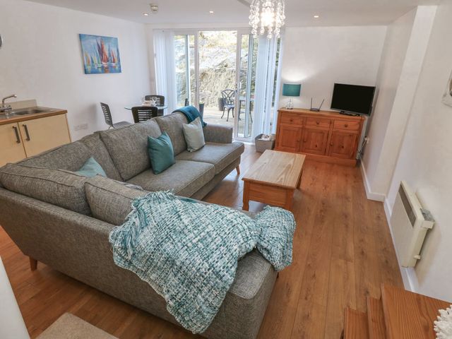 A living room with a gray sectional sofa a wooden coffee table a wooden cabinet with a TV and lamp and a dining area near glass doors at Beach Court Hideaway in Trearddur Bay