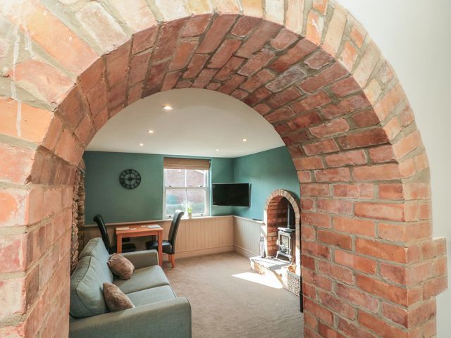 A living room with a green sofa two chairs a wooden table a wall clock a window a wall-mounted TV and a wood stove in a brick alcove at Top Holme Cottage in Newbald