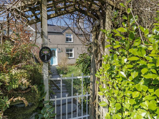 A pathway with stone slabs leading to a house with a blue door seen through a wooden arch with a white gate and foliage at 1 Aberbeinw in Dolwyddelan