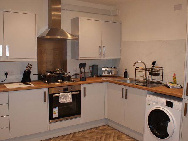A kitchen with white cabinets a washing machine a built in oven a stove with pots a drying rack on the sink and a countertop with small appliances at The Newborough sleeps 8 Apartments Scarborough