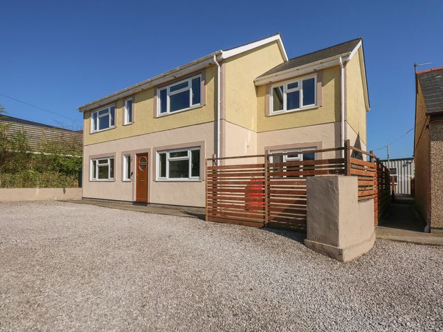 A two-story house with yellow and beige walls with a brown wooden door and multiple windows with a gravel driveway and a wooden fenced area at Criw Newydd in Bryncroes near Sarn Meyllteyrn