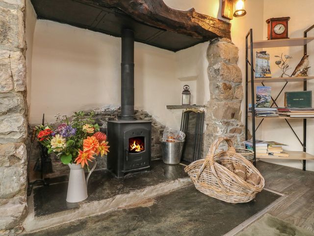 A fireplace with a wood burning stove and a basket of logs next to a metal bucket and a vase of flowers at Tea Cosy Cottage in Marhamchurch
