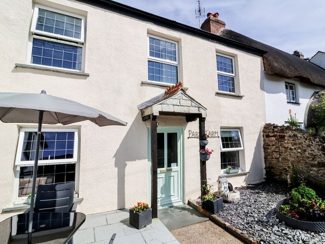 The exterior of a white house with a light green door a patio umbrella a chair and flowers at Tea Cosy Cottage in Marhamchurch
