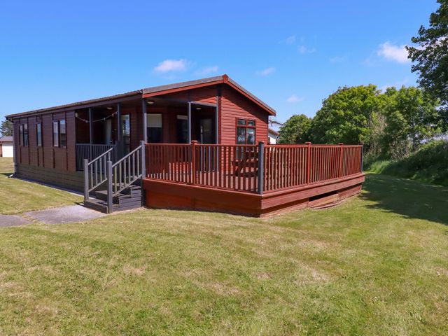 A wooden cabin with a fenced deck and steps on a lawn with trees in the background at Lodge 9- Lamorna in Tregolds near St Merryn