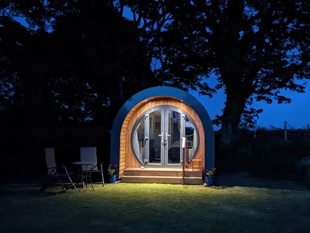 A small round wooden pod with glass double doors lit at night with two chairs and a table outside at Florence Pod in Pembroke