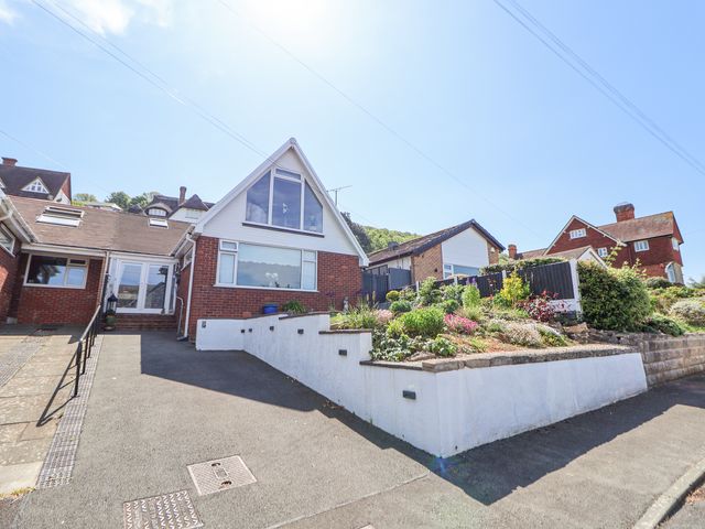 A brick house with a large window and sloped roof next to a garden and driveway at 67 Pendre Avenue in Prestatyn