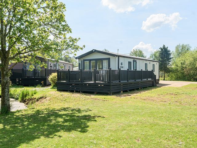 A mobile home with a black deck railing surrounded by grass and trees at Aquadrome Abode - Billing Aquadrome Holiday Park