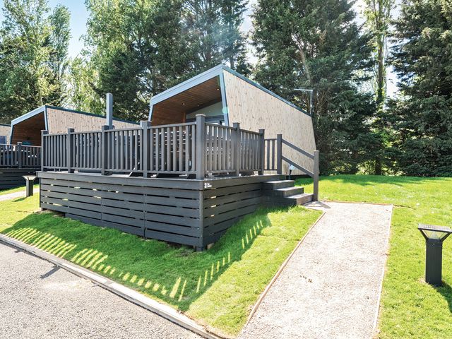 A wooden cabin with a deck and railing next to a gravel pathway in a forested area at Billing Aquadrome Holiday Park