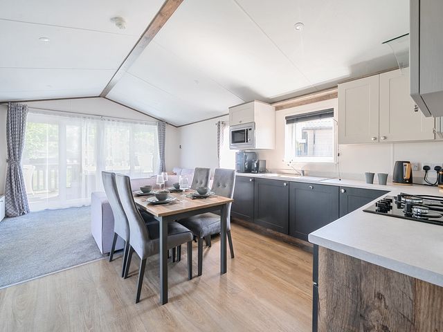A kitchen and dining area with a wooden dining table set for four and a living room with large windows at Wave Whisperers - Hayling Island in Hayling Island Holiday Park
