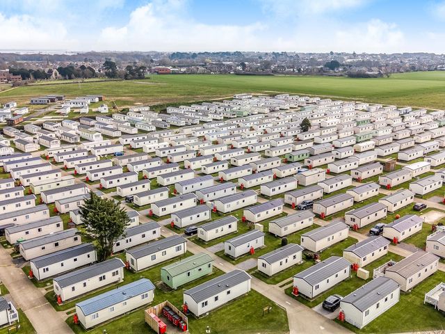 An aerial view of numerous caravans arranged in rows with green fields and houses in the background at Dune Delight in Hayling Island Holiday Park