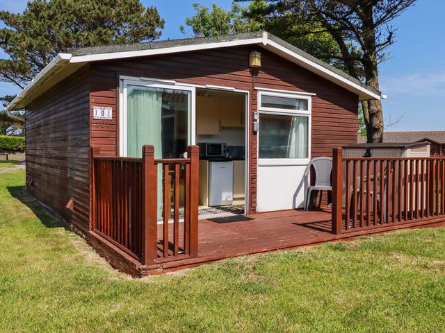 A small wooden cabin with a sliding glass door and a small porch with plastic chairs at 101 Atlantic Bays Holiday Park in Padstow