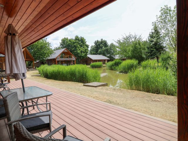 An outdoor area with a deck, table and chairs overlooking a pond at Sunnyside Lodge Thorpe-On-The-Hill