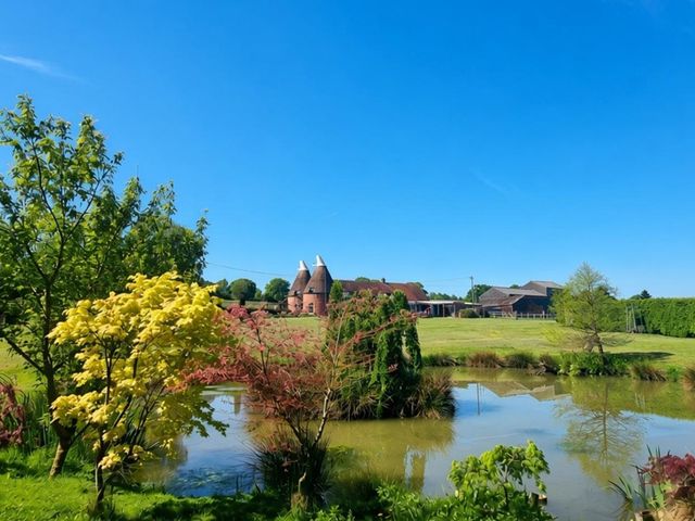 A house by a pond with trees in the foreground at Sharvels Oast House - Pool, Tennis, Luxury Home, Peasmarsh