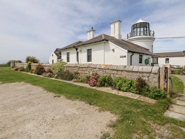 A white building with a stone wall and flowers in front and a lighthouse behind at Old Higher Lighthouse Stopes Cottage in Portland Bill