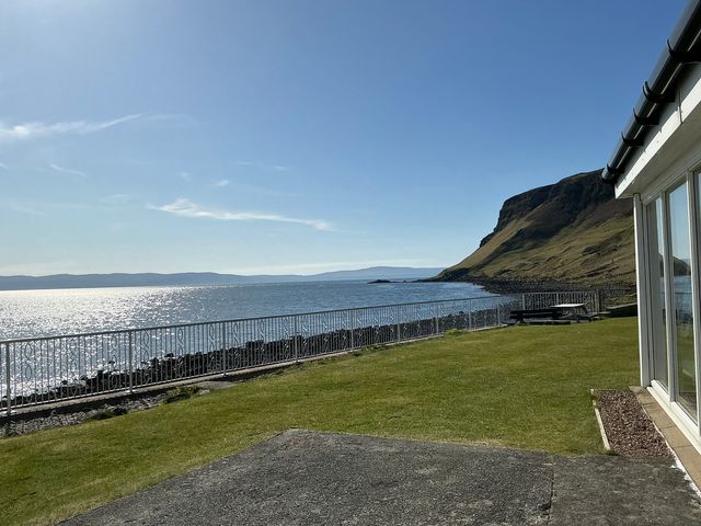 A grassy yard overlooking a body of water with a metal fence and a rocky hill near Jubilee Cottage in Uig