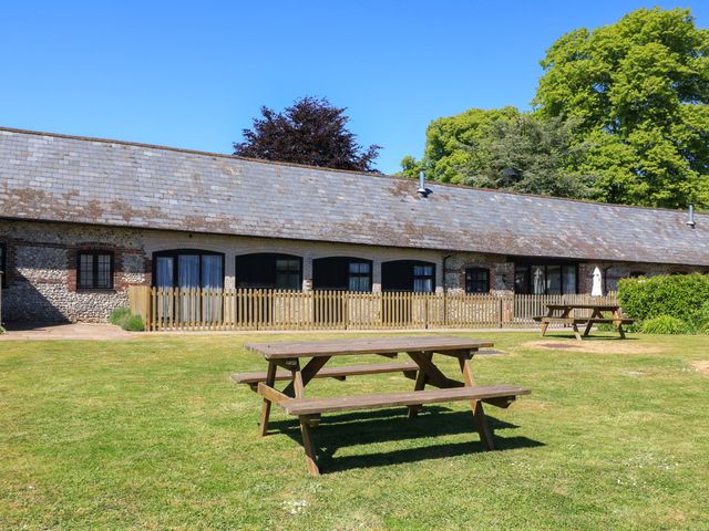 A grassy outdoor area with wooden picnic benches and a building with a tiled roof and fenced patio at The Old Stables in Winterborne Stickland