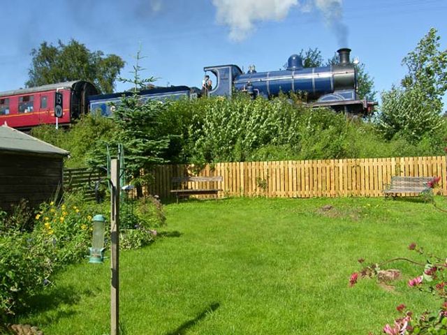 A red and blue steam train passing behind a garden with a wooden fence and benches at Steam Cottage in Aviemore