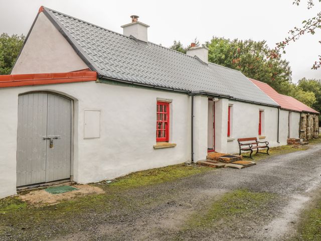 A white cottage with a gray tiled roof red window frames and a wooden bench outside at Willowbrook Cottage in Askill County Leitrim