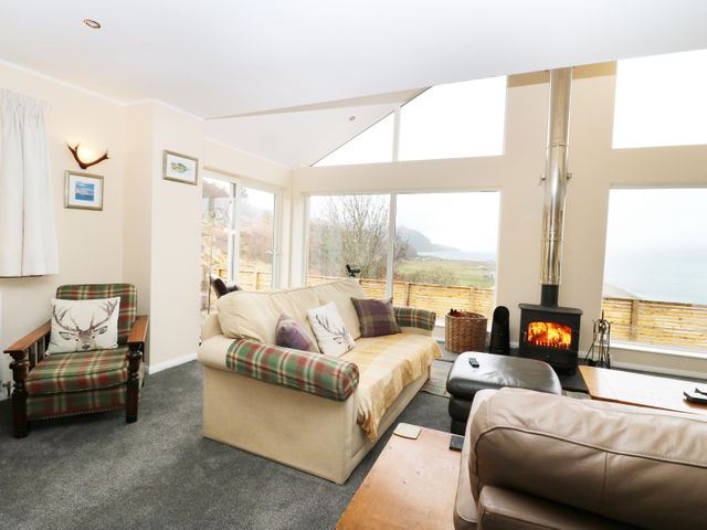 A living room with a sofa armchair and wood burning stove with large windows showing an outdoor view at Torgorm in Glenelg