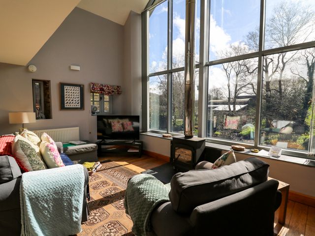 A living room with armchairs a wall-mounted tv and a wood-burning stove by large windows at The Barn at Glanoer in Hundred House near Builth Wells
