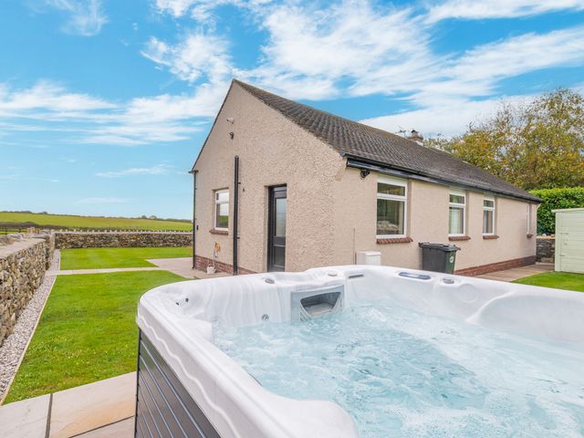 A hot tub in a backyard next to a single-story house with a stone wall and grassy area at Colt Park Cottage in Ulverston