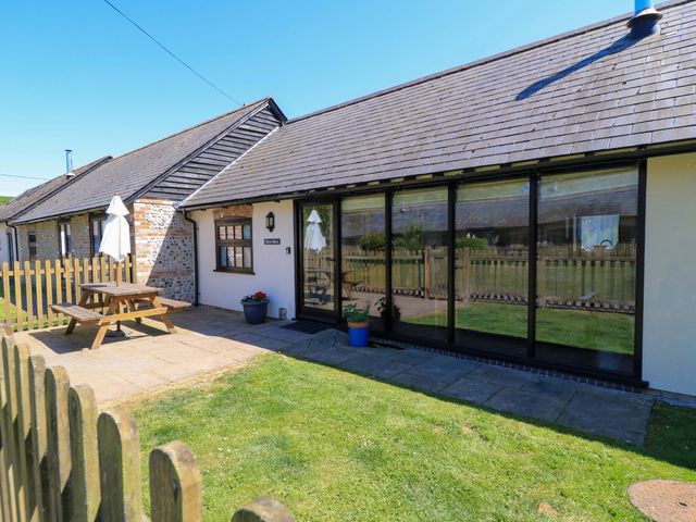 An exterior view of a cottage with a wooden picnic table and umbrella on a paved patio at New Barn in Winterborne Stickland