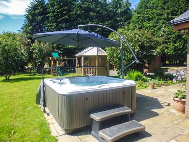 An outdoor patio area with a hot tub under an umbrella and garden play equipment in the background at Tynddol Bungalow in Llanbadarn-Fynydd near Llanbister