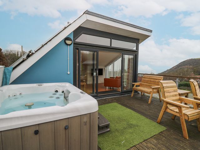 An outdoor deck with a hot tub wooden chairs and a bench next to a building with glass sliding doors at Ty Henri in Tresaith