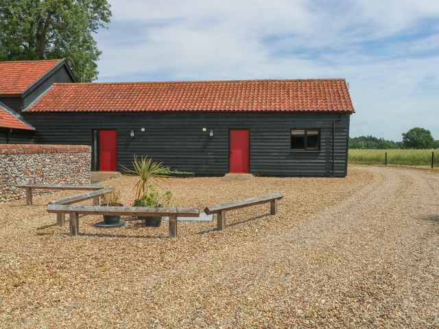 A black wooden building with red doors and a gravel driveway with wooden benches and plants at Snowy Owl Barn in Shipdham