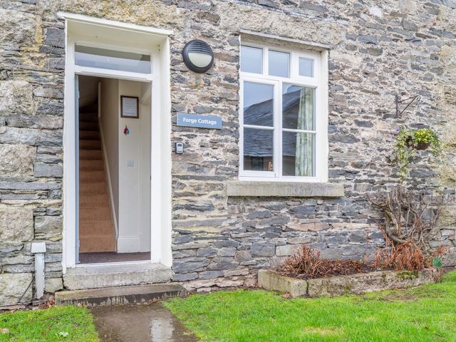 Stone exterior wall of a cottage with an open door showing stairs and a window with a flower basket hanging beside it at Forge Cottage in Graythwaite near Hawkshead