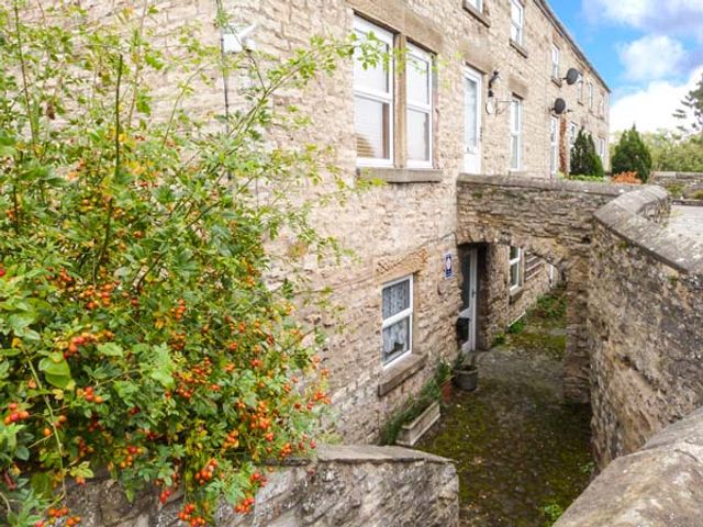 A stone building with windows and a door under a stone archway with plants growing nearby at 2A Bridge Apartments in Masham near Ripon