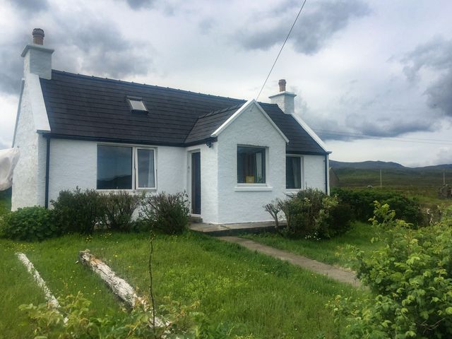 A white cottage with black roof and chimney in a grassy area with shrubs at Ambers Cottage in Staffin