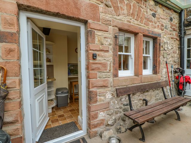 An open door showing a kitchen with a trash bin a wooden bench outside against a stone wall with two windows and cleaning tools nearby at Blacksmith Cottage in Appleby-in-Westmorland