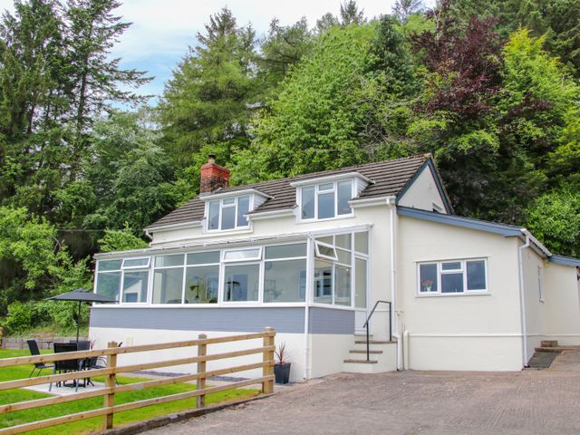 A white house with large windows and a small patio area with a table and umbrella surrounded by trees at Caesamson in Caersws