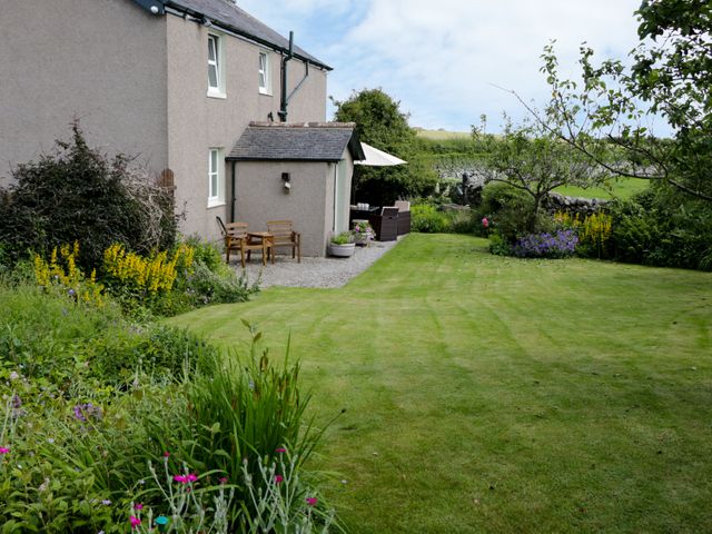 A garden with lawn, flower beds, trees, wooden chairs, and a table with umbrella outside a house at Waingate Cottage in Flookburgh