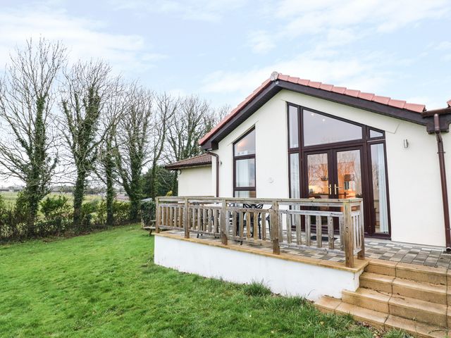 A house with a tiled roof brown framed windows and a wooden deck with a patio table and chairs surrounded by grass and trees at Keepers Cottage in Sidmouth