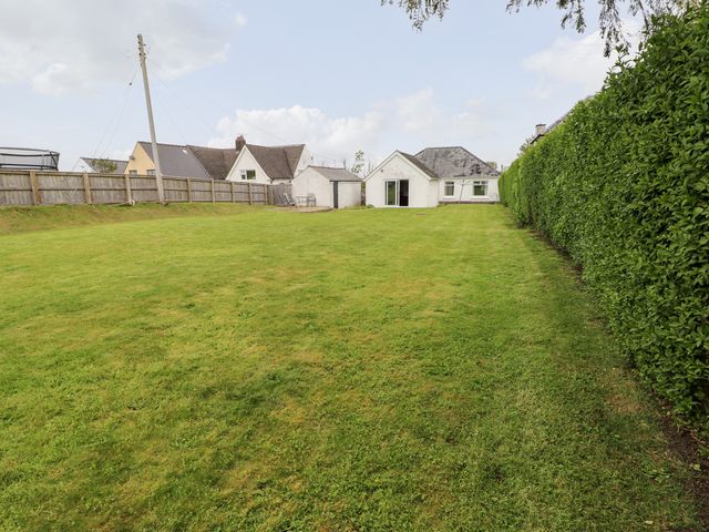A large grassy backyard with a white house and two sheds bordered by a wooden fence and green hedge at White Thorns in Haverfordwest