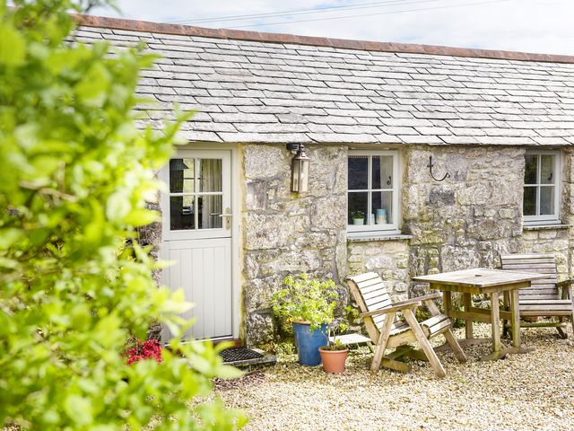 A stone cottage exterior with a white door wooden chairs and a table on a gravel patio at The Snug in Rescorla near St Austell