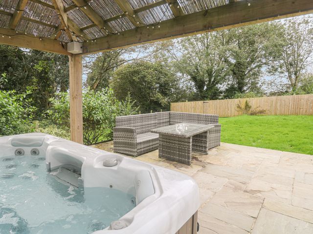 An outdoor patio with a hot tub under a wooden pergola and wicker seating with a table on a stone floor and grass and trees in the background at Kelyow in St Day