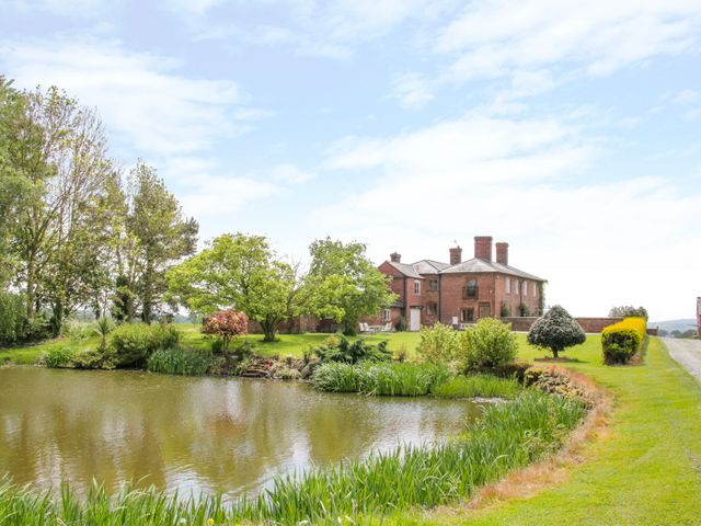 A large brick house with chimney stacks near a pond surrounded by grass and trees at The Manor House at Kenwick Lodge in Cockshutt near Ellesmere